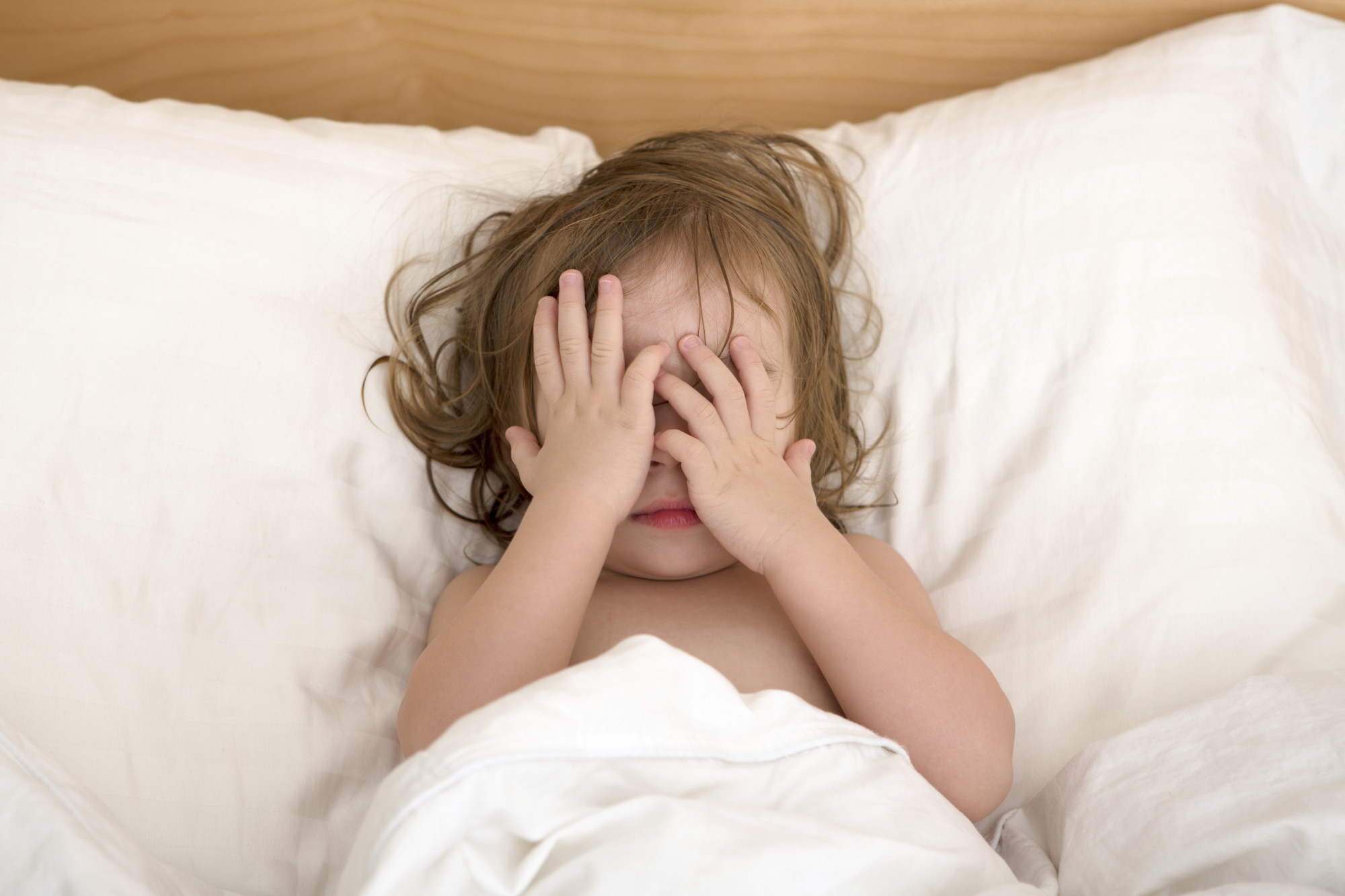A little girl laying on a bed with white bed sheets with both hands covering her face