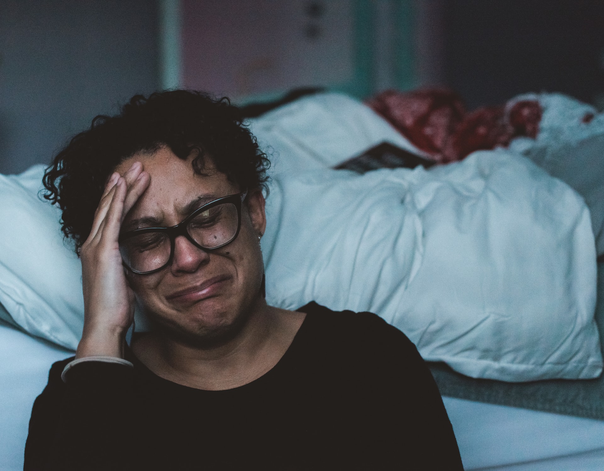 A woman wearing an eyeglass sitting on the floor beside her bed crying