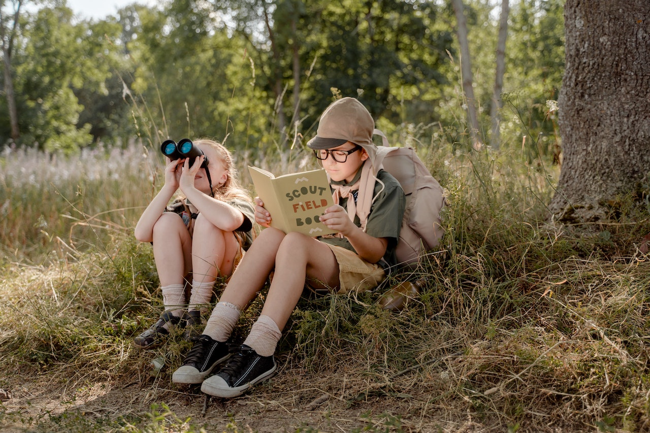 Blonde Girl Looking at the Binoculars While The Boy Is Reading A Scout Field Book