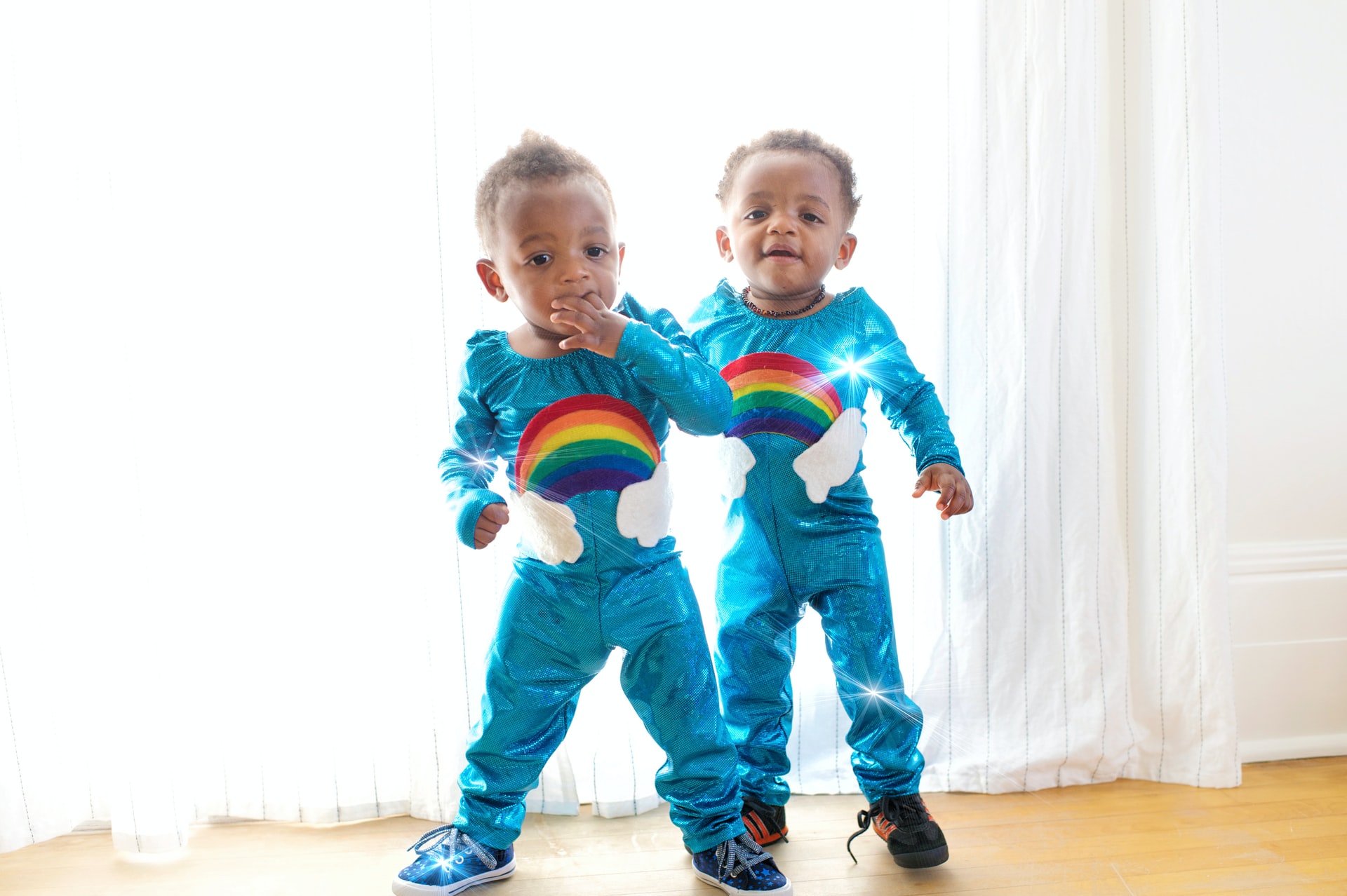 Male black twins wearing matching blue onesies with rainbow standing side by side