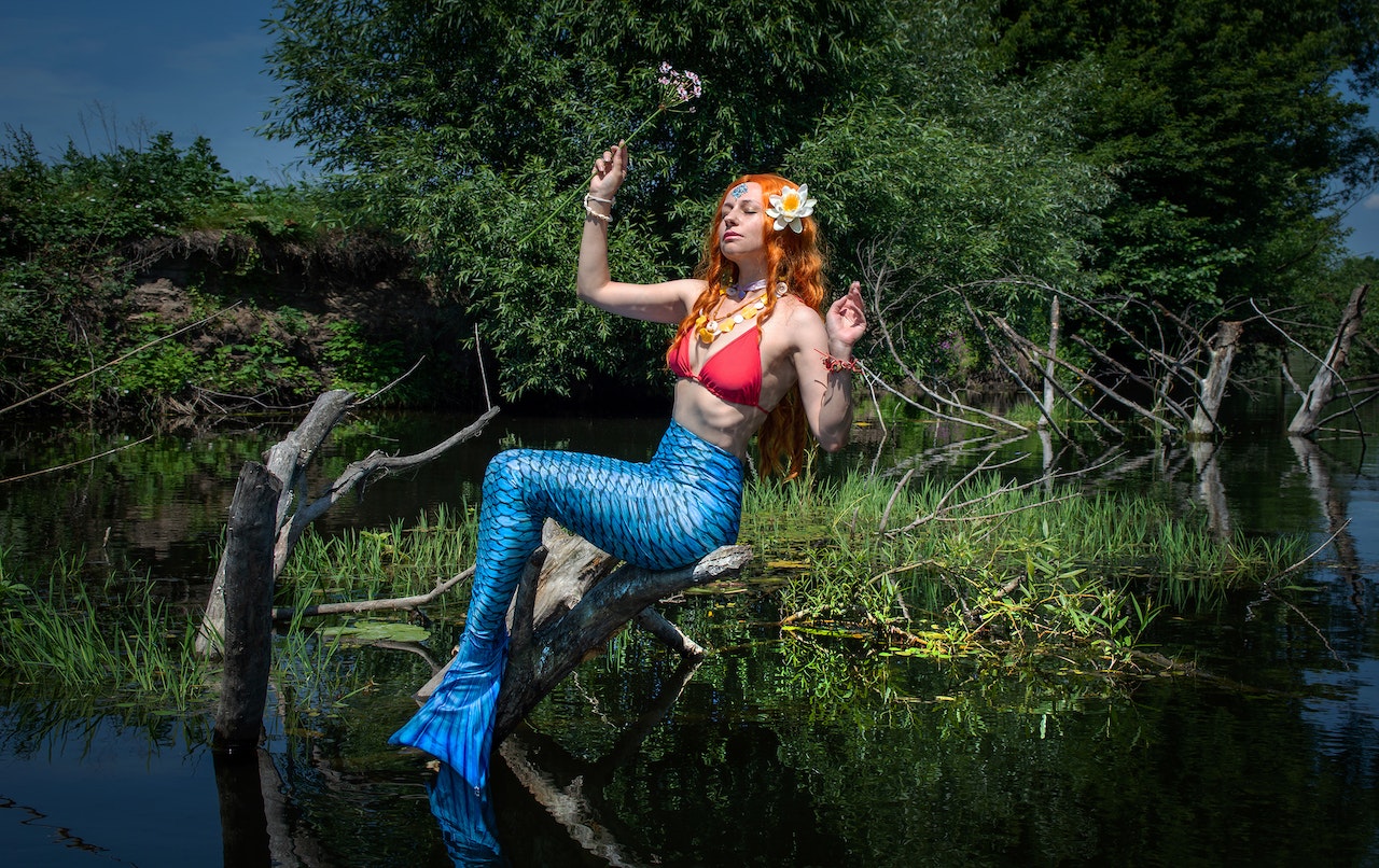 A Beautiful Mermaid Holding A Flower while Sitting On A Wooden Log On A River