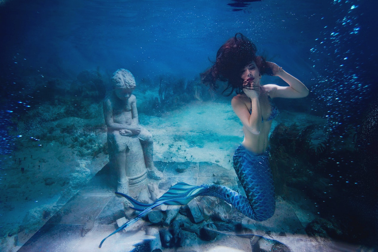 A Mermaid Underwater With A Statue Of A Child