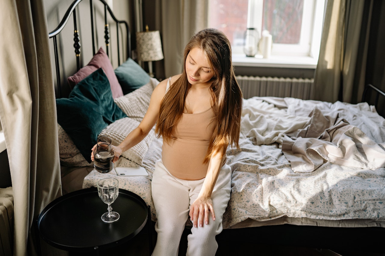 Pregnant Woman Pouring Liquid on Clear Drinking Glass While Sitting On The Bed With Messy Hair