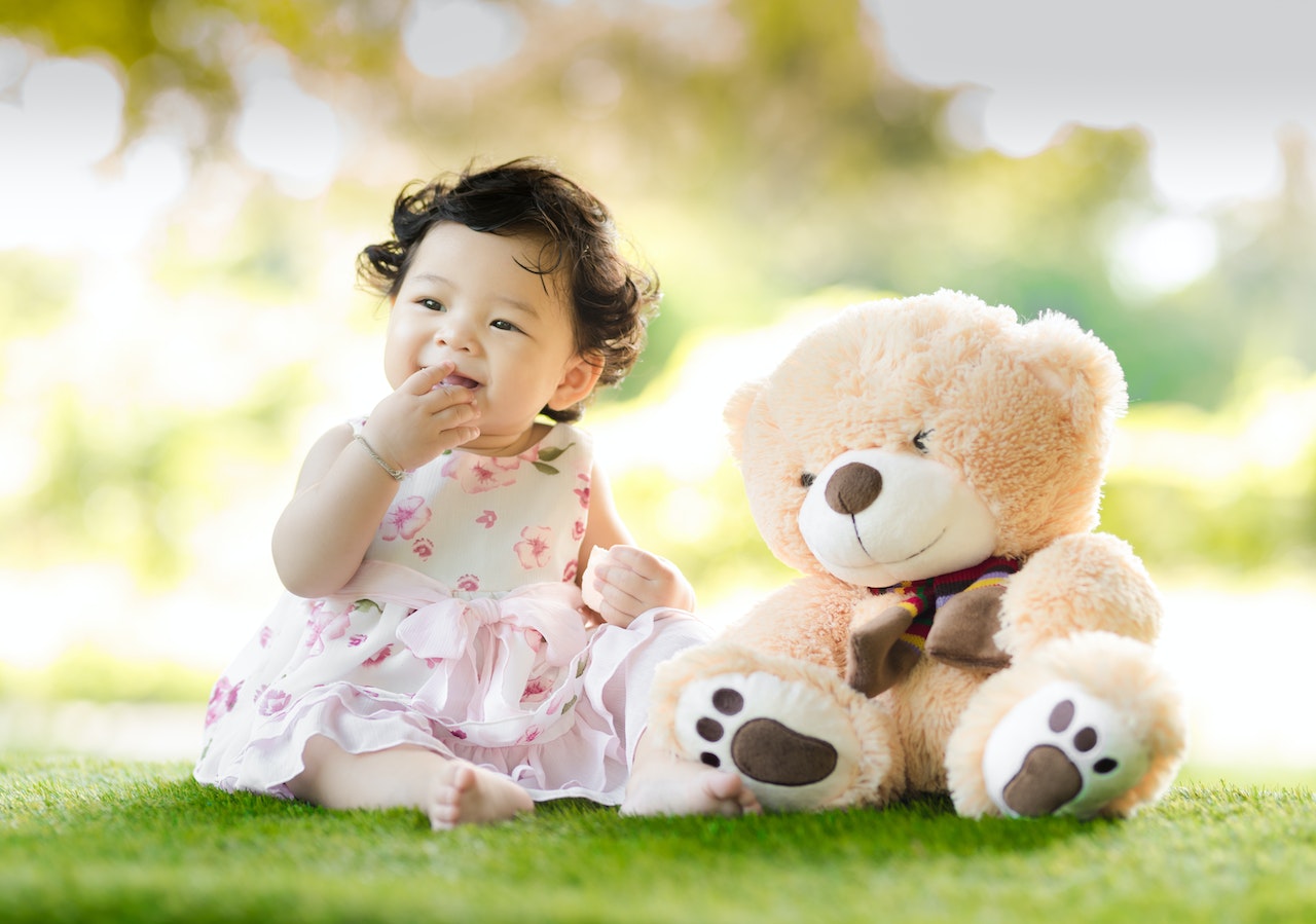 Baby Girl Sitting on Green Grass Beside A Bear Plush Toy at Daytime