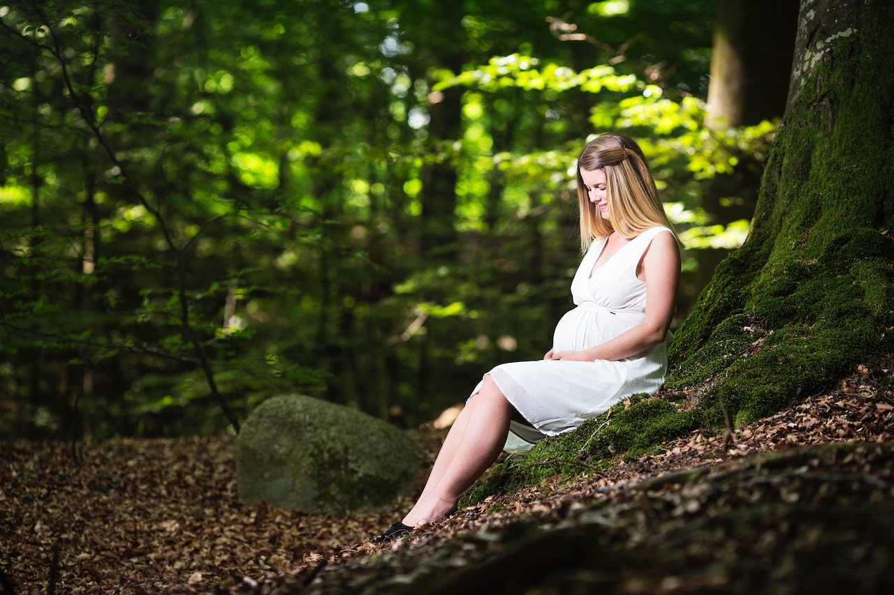 Pregnant Woman in a White Dress Sitting On a Tree Trunk