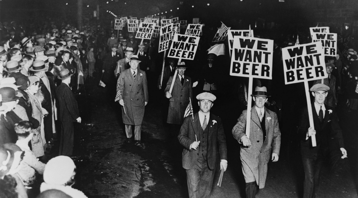 A crowd of people with some standing and some walking holding a placard with the word "we want beer" written on it