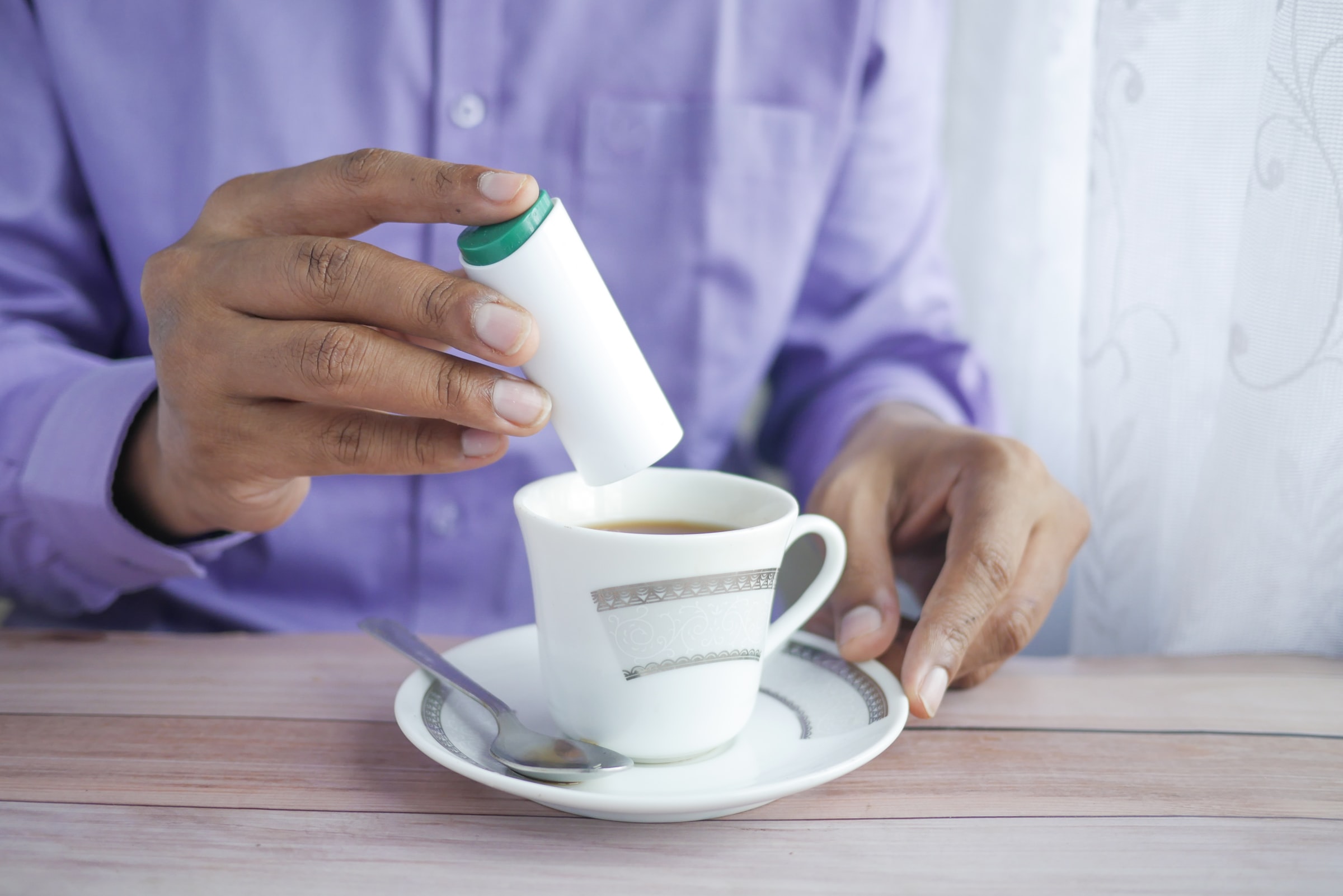 A black man wearing a purple shirt pouring sugar into his cup of tea