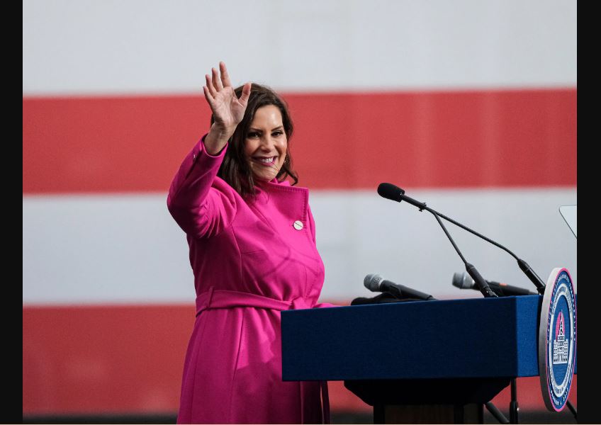 Gretchen Whitmer wearing a pink jacket while standing on a podium with several microphones as she smiles and waves