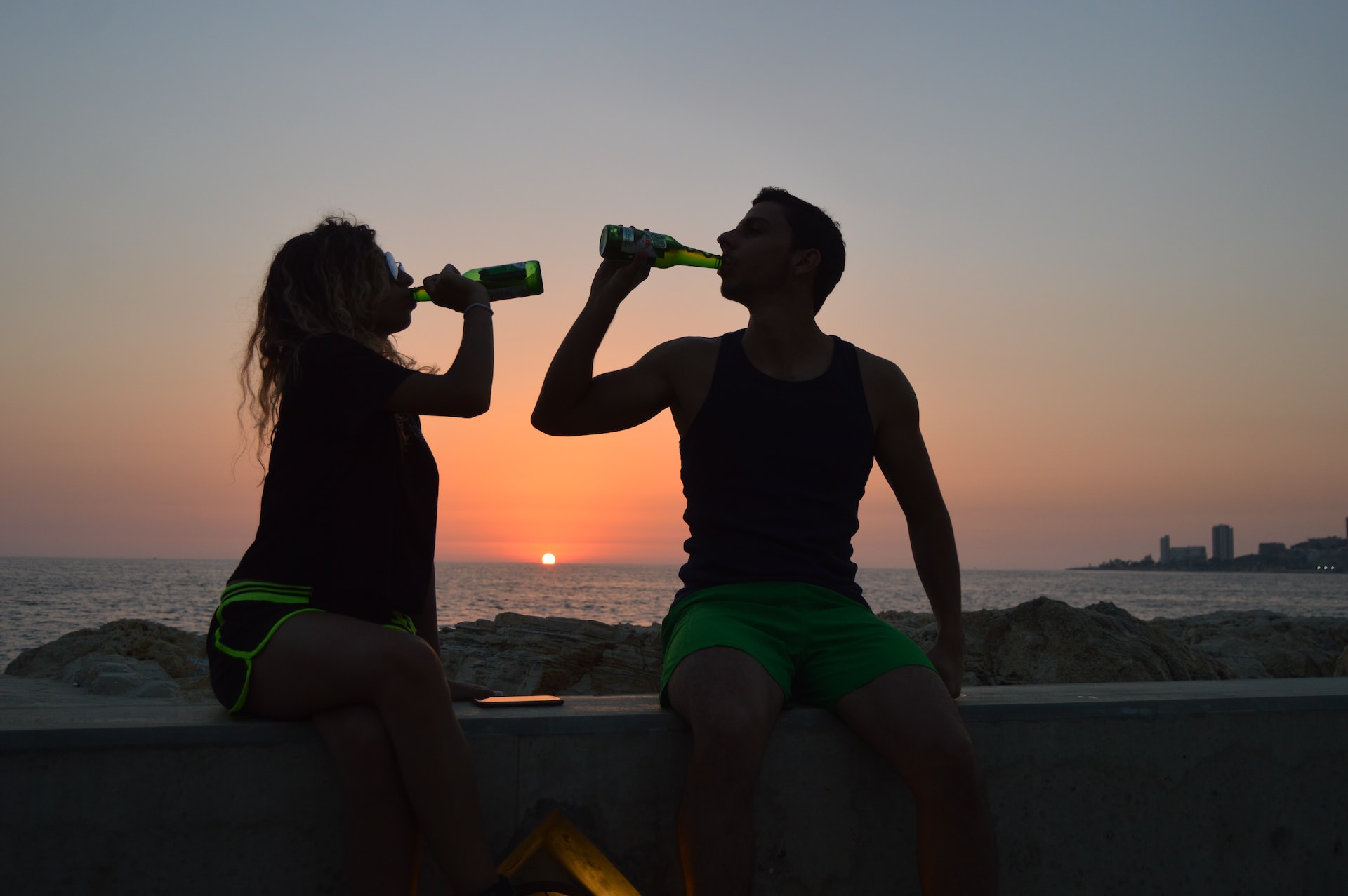 A male and female adult seated near the sea drinking dealcoholized wine straight from the bottle at sunset