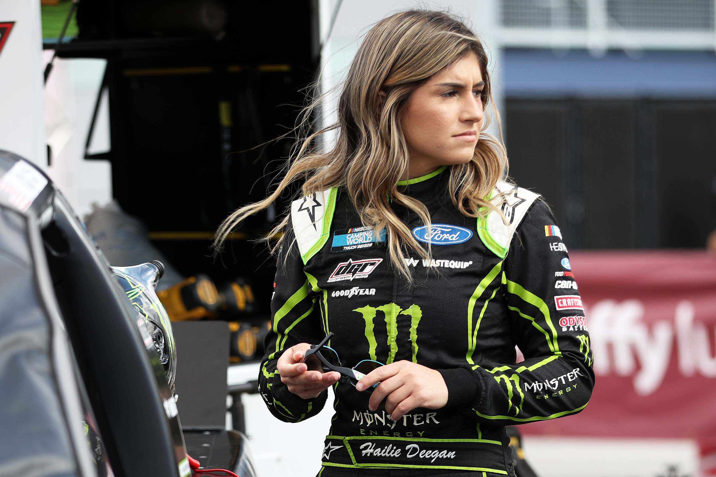 Hailie Deegan wearing her racing attire while holding a black eye glass and standing beside her car