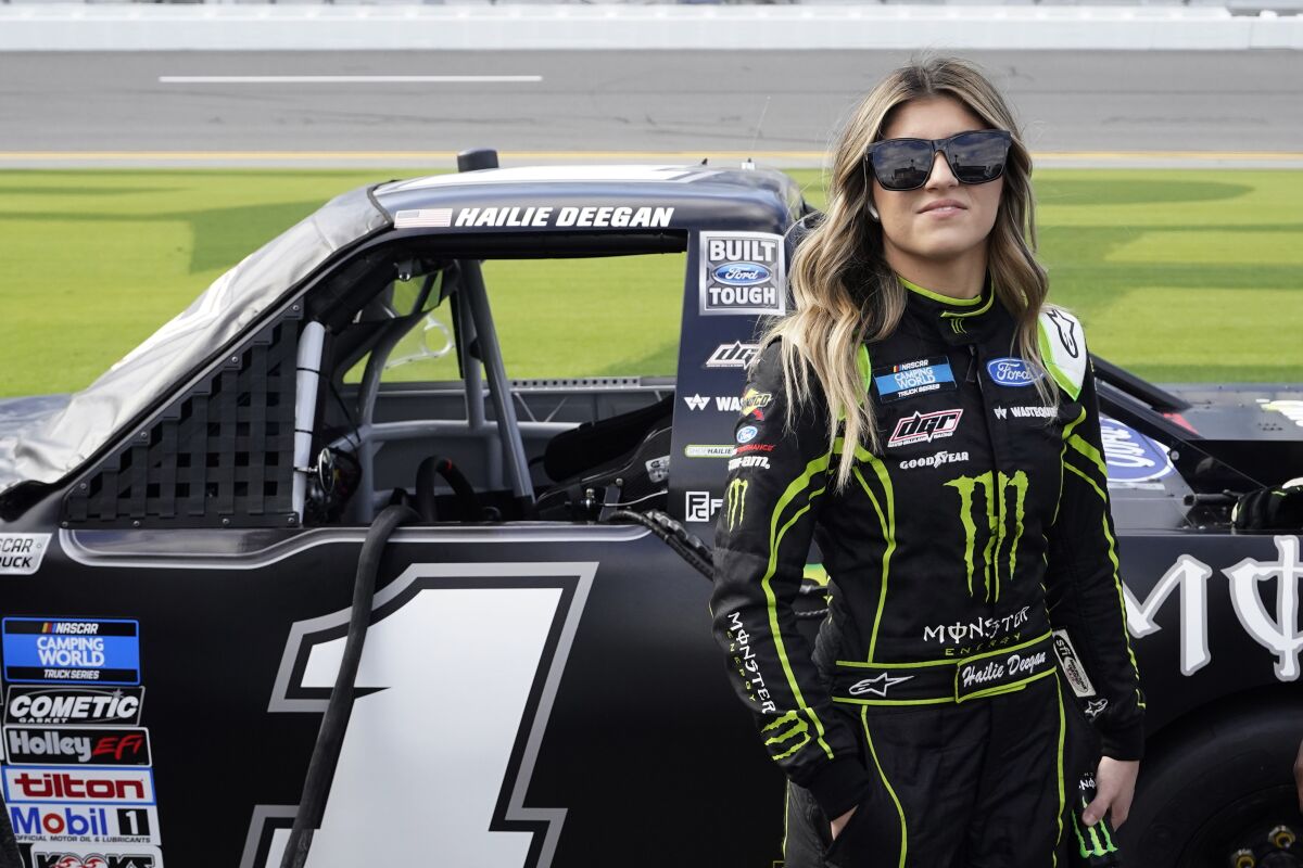 Hailie Deegan wearing her racing attire and a black eye glass as she stands beside her car