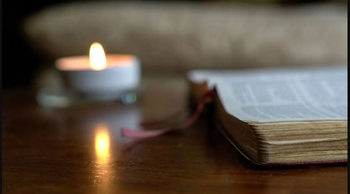 An open bible sitting beside a lit white candle on a wooden surface