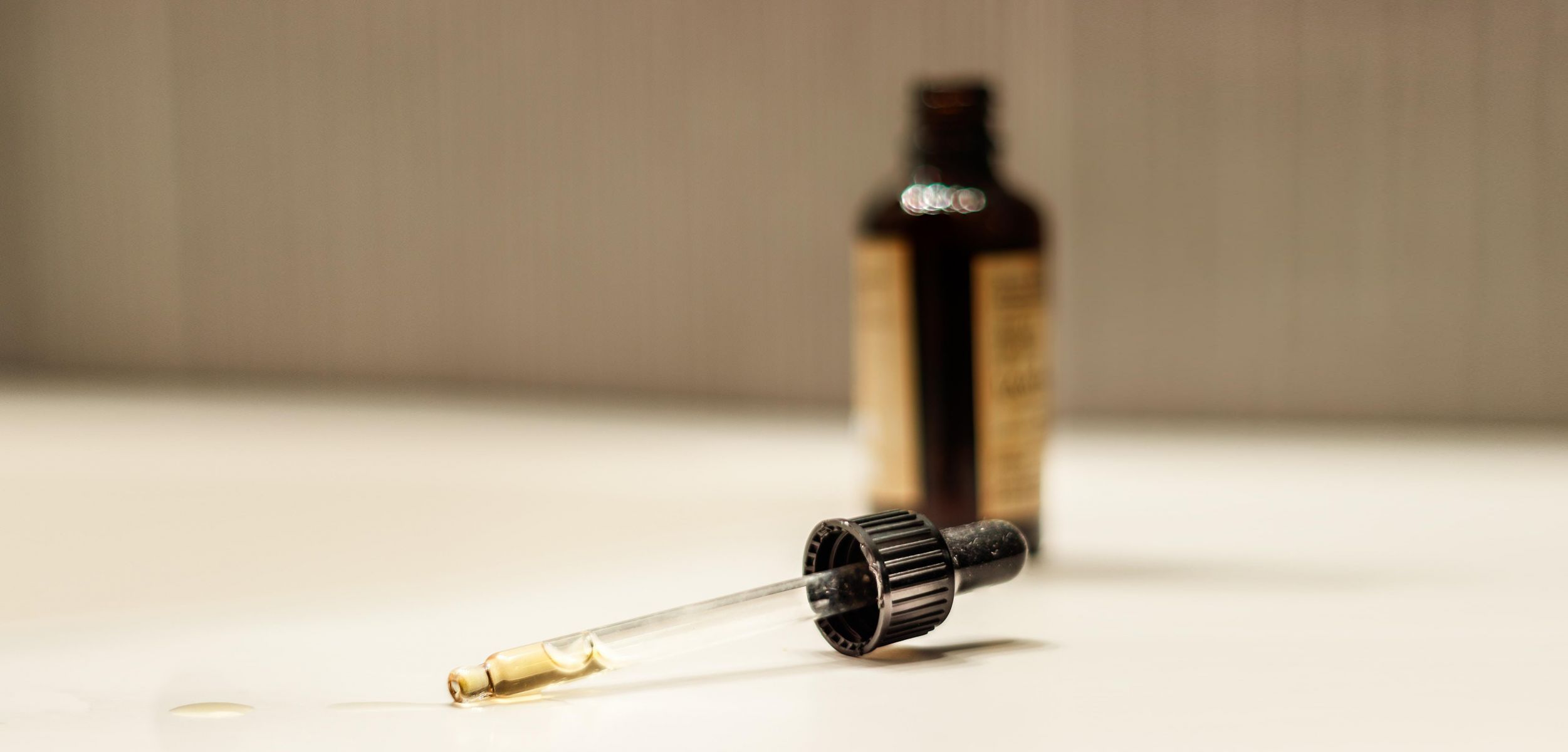 A CBD oil tincture dropper with few drops of CBD oil in it on a blurry background of an opened brown CBD oil tincture bottle