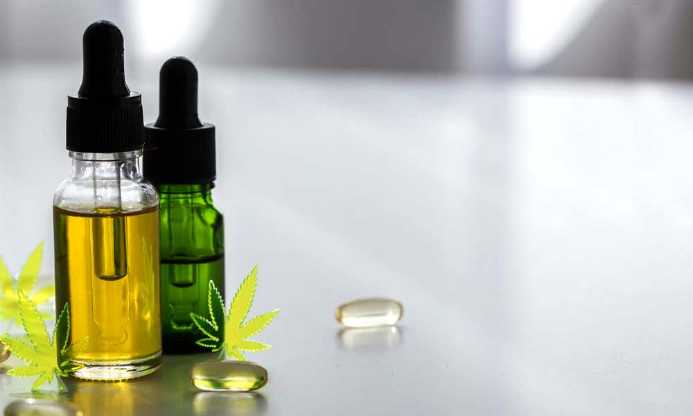 A closed transparent tinture bottle filled with CBD oil and a closed brown tincture bottle filled with CBD oil sitting beside two transparent CBD capsules