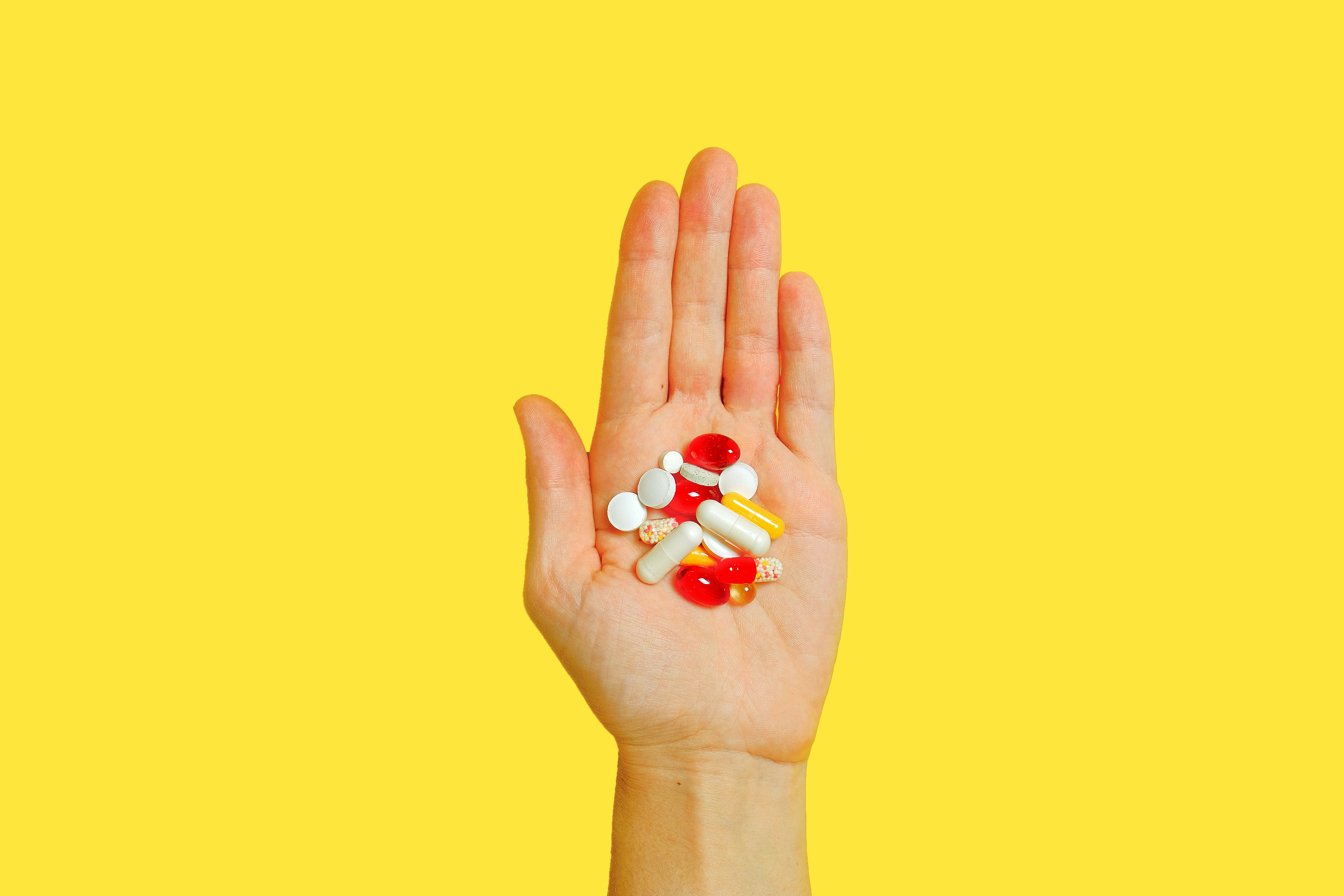 A Caucasian opened palm holding drug tablets of different sizes, shapes, and color on a yellow background