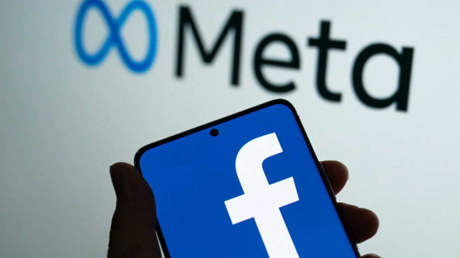 A hand holding a phone showing the Facebook logo with the Meta logo in the background