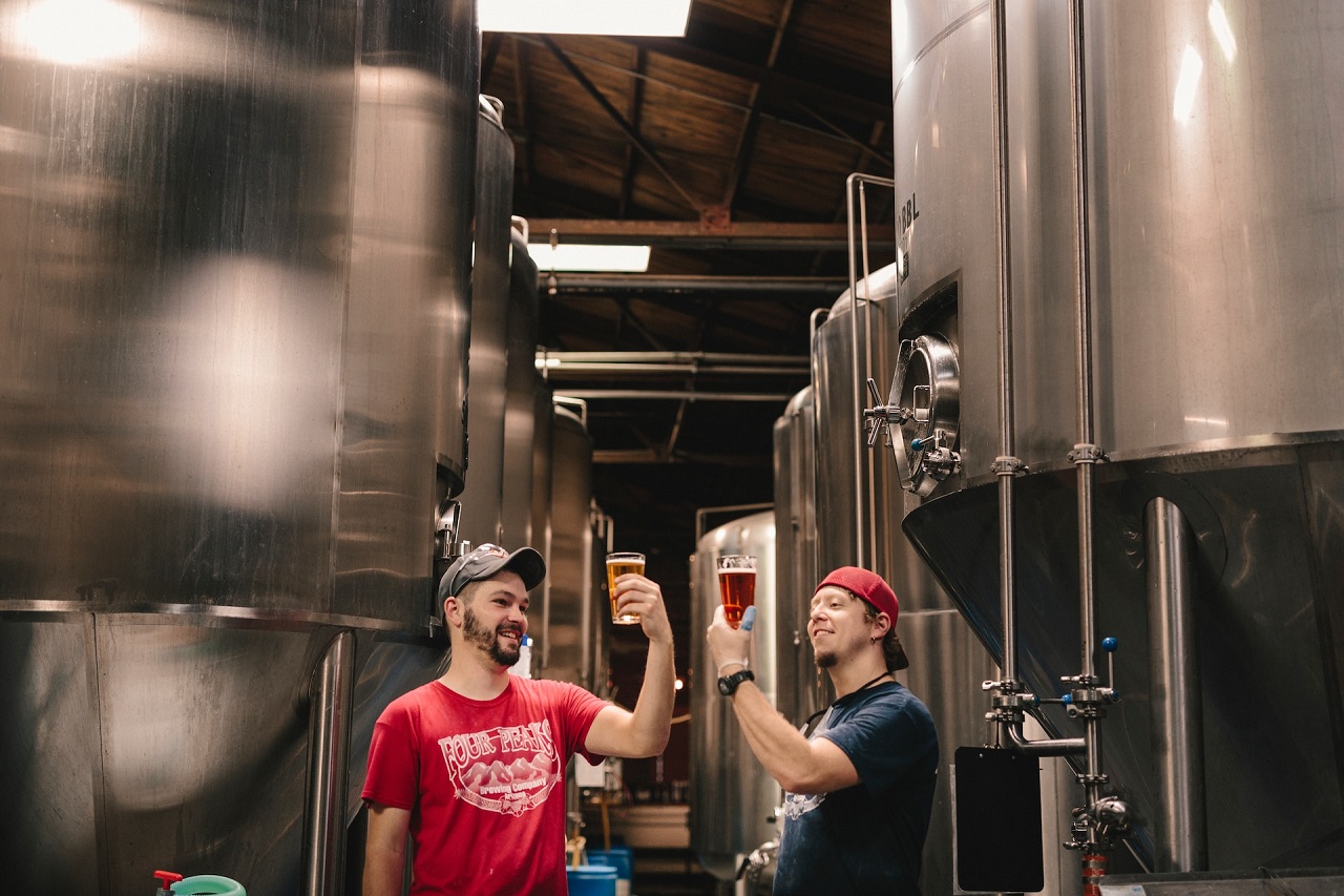 Two men in T-shirt and caps standing near cylindrical tanks and raising and looking at a glass of beer