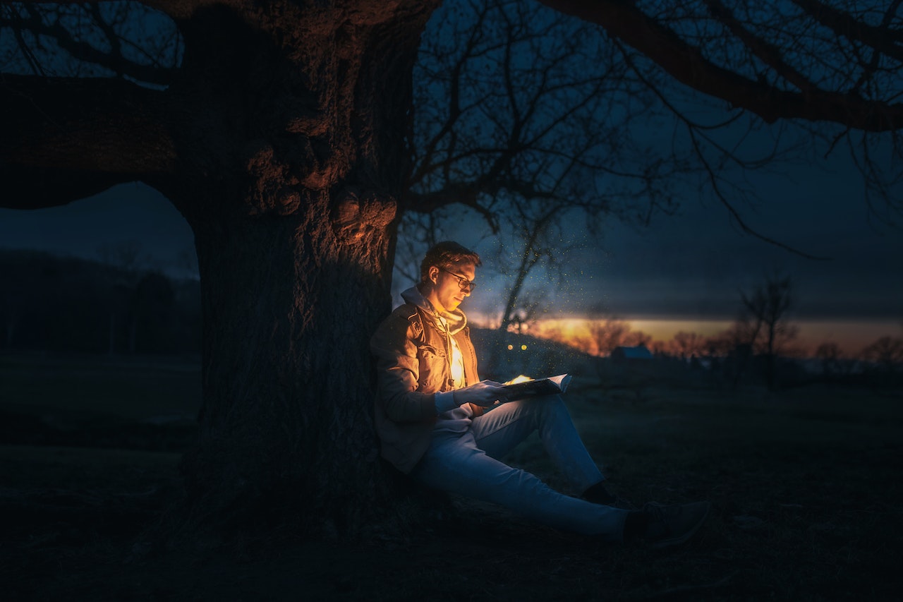 Man Sitting Under A Tree Reading A Magical Speels Book during Night Time