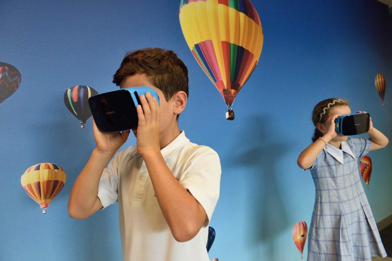 Boy and girl using blue virtual reality goggles in a room with wallpaper  and hot air balloons as design