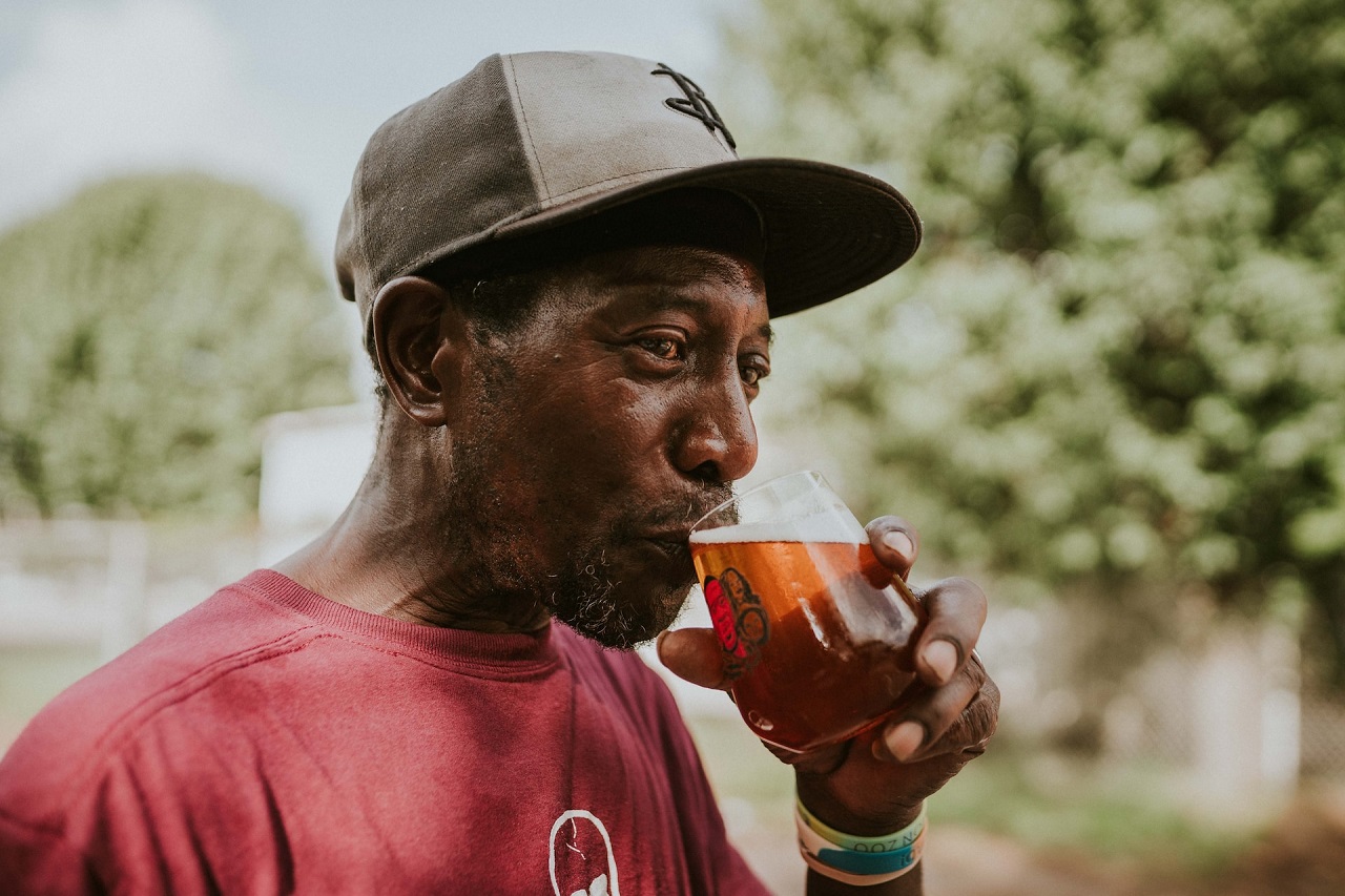 A black middle-aged man in a cap and red T-shirt drinking dealcoholized beer from a short stout glass