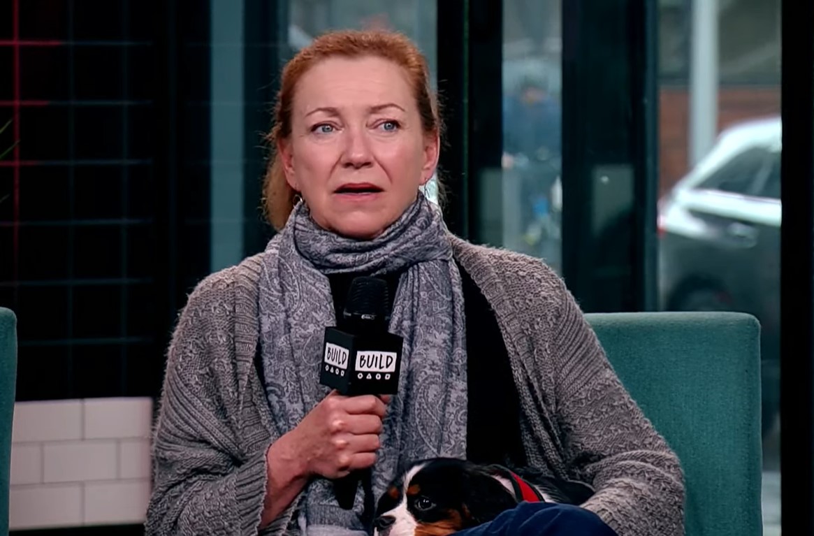 Julie White in gray sweater and giant dark scarf around her neck holding a mic and a puppy on her lap