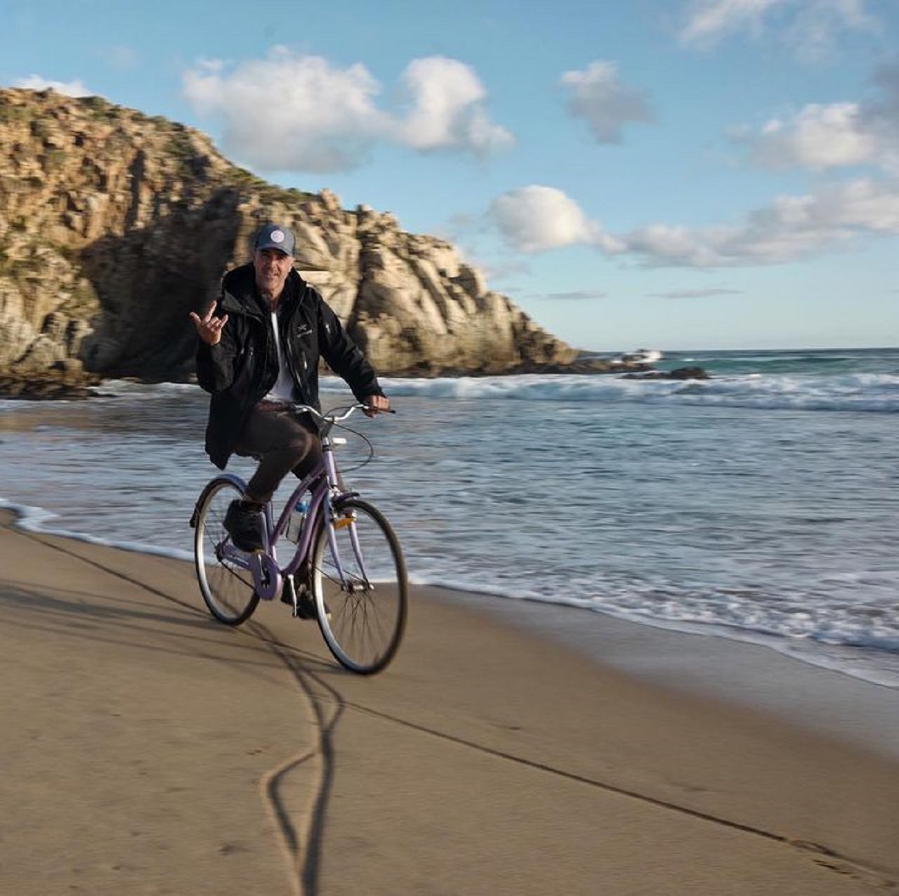 Jeff Pangman in a jacket and cap cycling along the beach and making the rock-and-roll hand sign