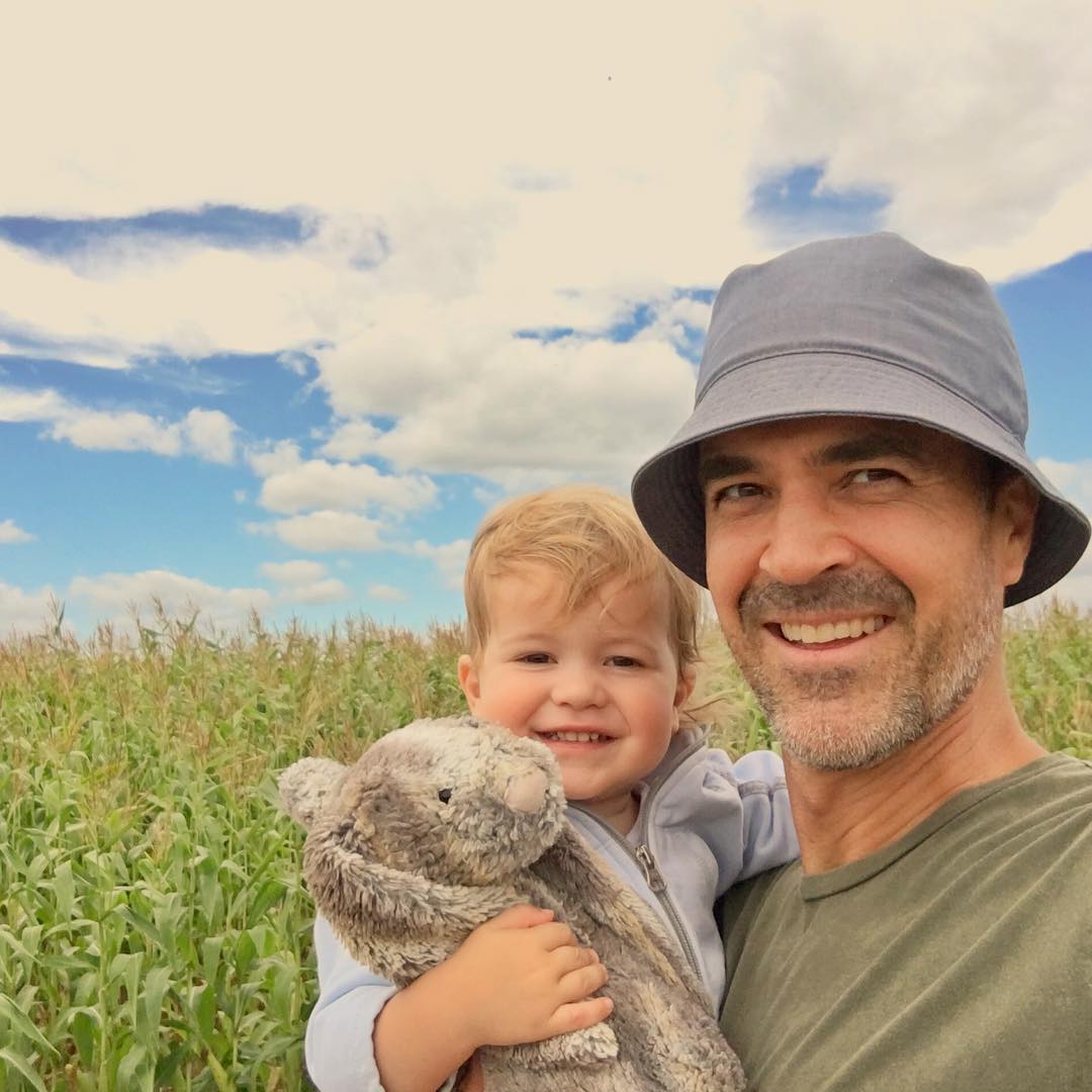 Jeff Pangman in bucket hat carrying his toddler son holding a rabbit stuffed toy on a field with tall green crops